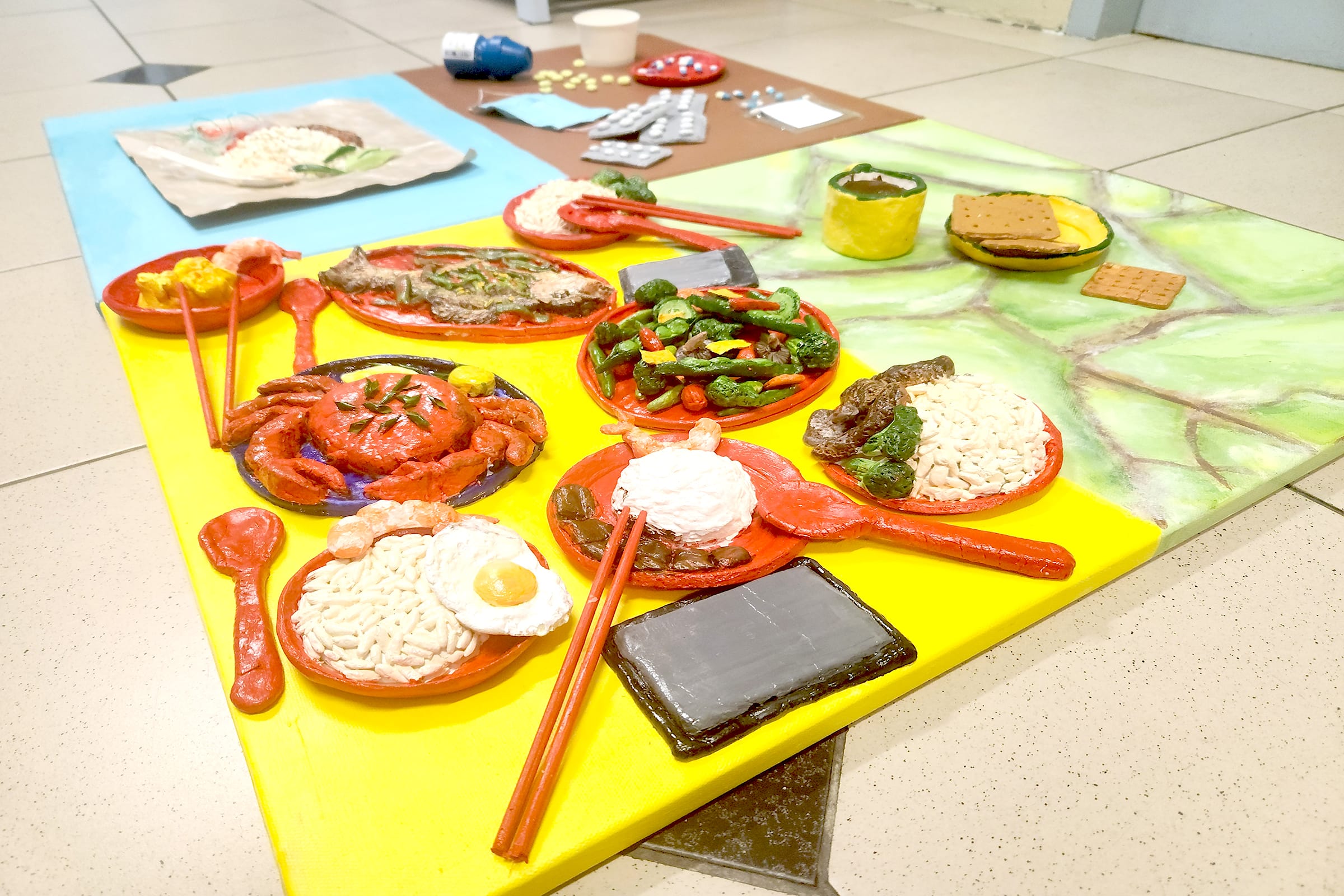 Colorful clay or painted sculpture of Asian meal dishes with chopsticks, crab, rice, and vegetables on a yellow board.
